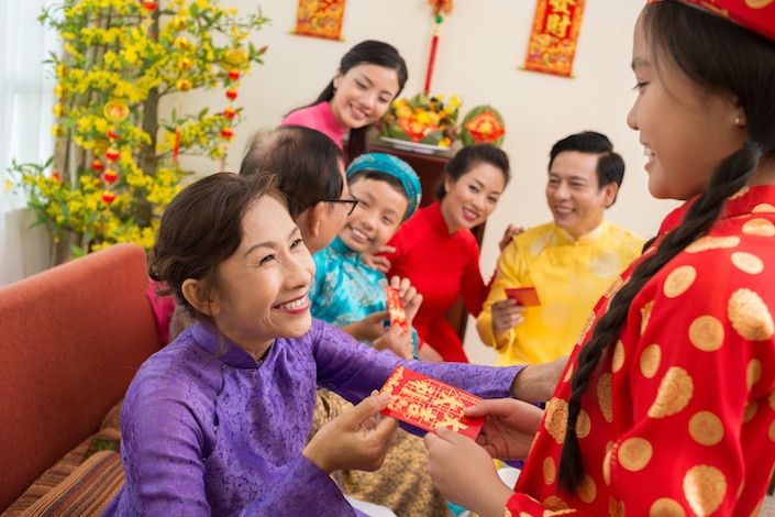 una mujer china mayor sentada con una camisa morada le da un hongbao a una joven china que está parada frente a ella y sonriendo