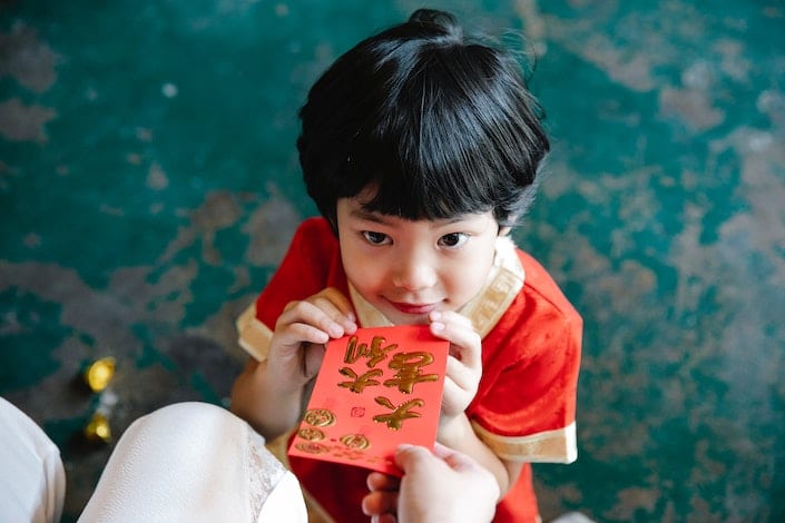 a little Chinese child with short black hair reaching for a Chinese hongbao being given to her by a person seated in front of her
