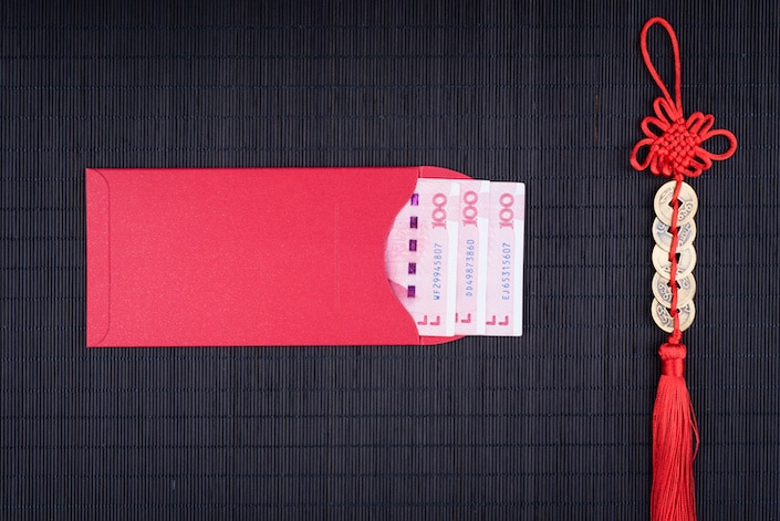 一个中国红包，上面贴着三张中国 100 元人民币的纸币，水平放置在一串圆形中国硬币旁边的黑色表面上