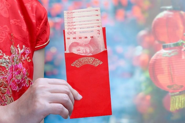 a woman's hand holding a Chinese red envelope with Chinese 100 RMB notes peeking out and Chinese lanterns in the background