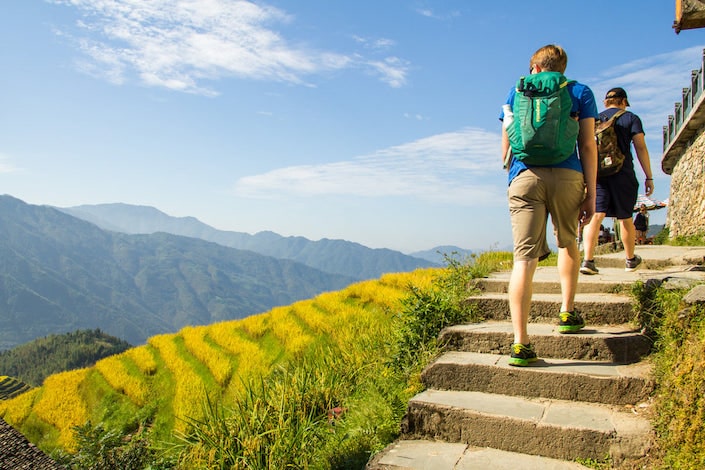 two young western men with their backs to the camera climbing stone stairs at the top of a mountain covered with terraces rice fields with other bare mountains rising in the distance