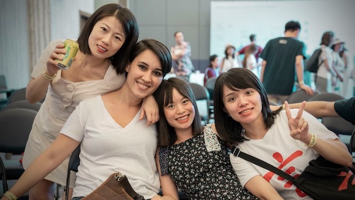 a group of four young women, three Chinese and one western, three of whom are sitting down and one of whom is standing up with a juice can in her hand, and all of whom have their arms around each other