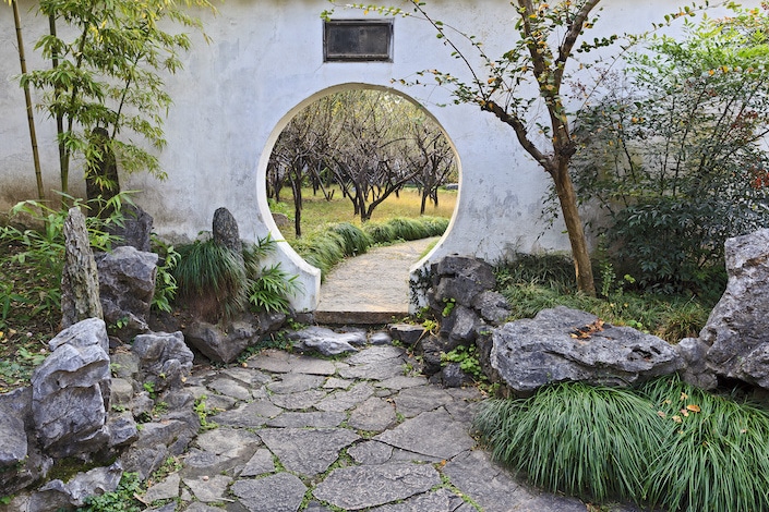 Pared blanca de jardín tradicional chino formal con orificio de puerta redondo de bambú y piedras decorativas junto a árboles frutales