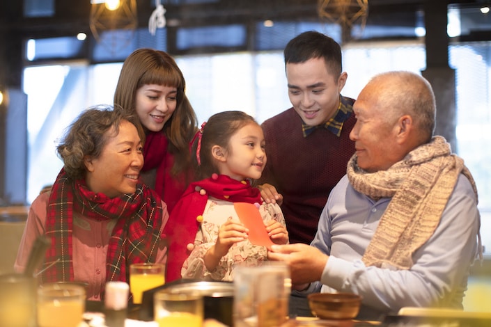 a picture of a three generation Chinese family including a little girl, two grandparents and a mother and father; the granddaughter is receiving a red envelope from the grandfather