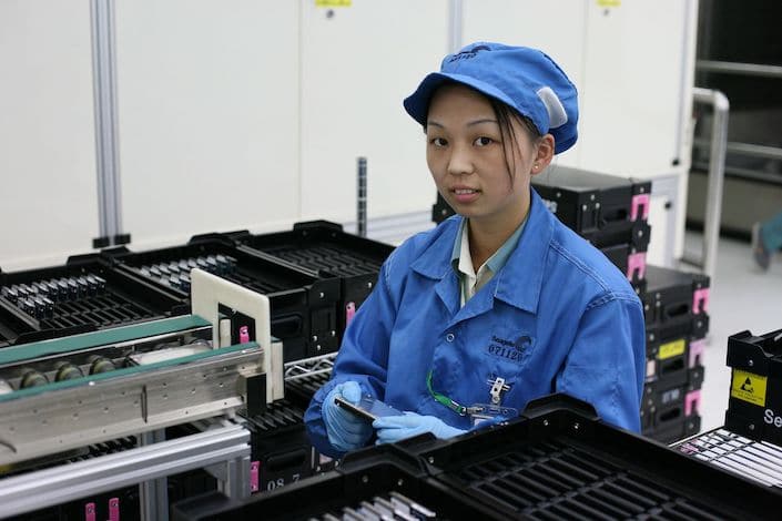 Una joven trabajadora de una fábrica china vestida con un uniforme azul y un sombrero de pie en una línea de montaje de productos electrónicos sosteniendo un teléfono celular con las manos enguantadas