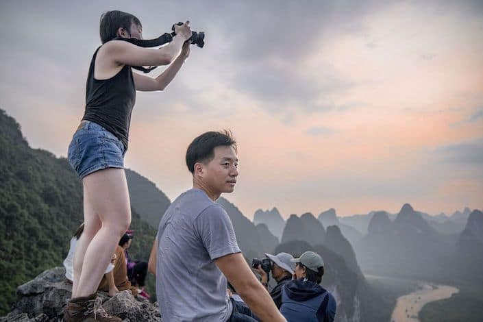 a chinese man sitting on a rock looking to the right of the photograph while a girl stands behind him holding up a camera taking a picture of the karst mountain peaks and river in front of them