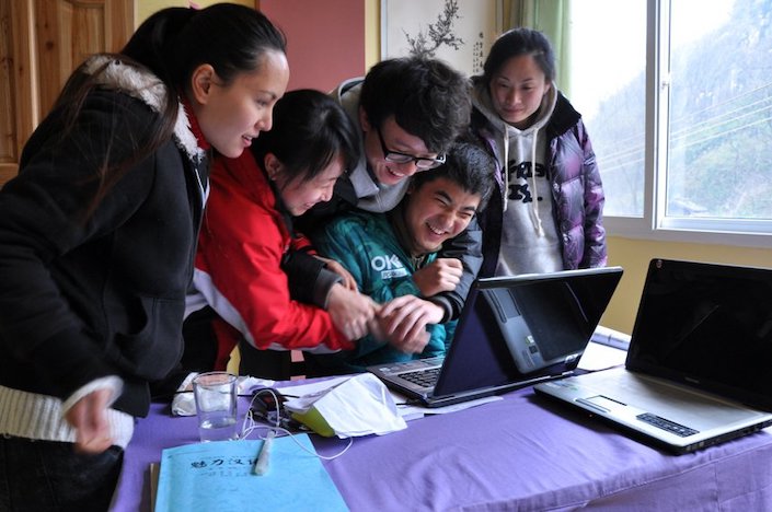 a group of five Chinese men and women hugging each other to keep warm while looking at a computer on a table next to a window