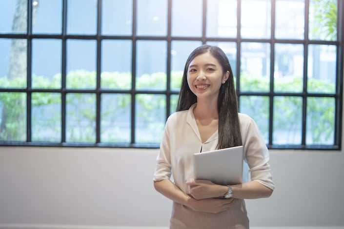 Employée d'assistance de bureau femme asiatique debout souriant tout en tenant une tablette avec les deux mains sur le ventre, vêtue d'une chemise à manches longues enroulée jusqu'au coude et d'une jupe rose.