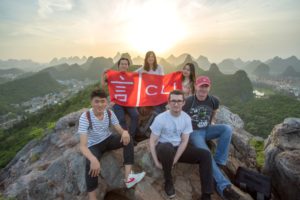 A group holds a CLI flag on a rocky peak above karst hills at sunset