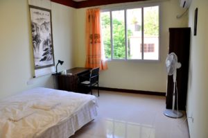 A simple bedroom with a desk, a fan and a scroll painting