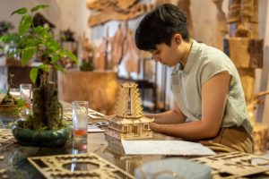 A person in a green shirt assembles a wooden pagoda model beside a bonsai