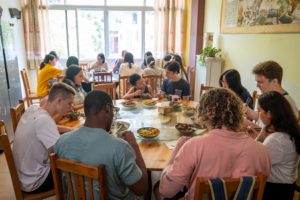 A big group eats around one large round table beneath a wall painting