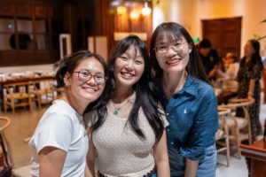 Three smiling women lean together for a photo inside a busy restaurant