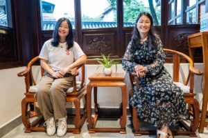 Two women sit in carved wooden chairs beside lattice windows