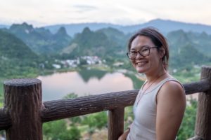 A smiling woman leans on a wooden railing above a lake and karst hills