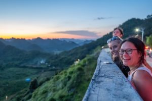 Friends smile from a stone wall overlooking green mountains at sunset