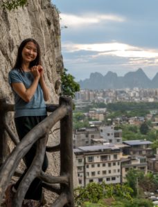 A woman stands on a rocky outcrop with the city and karst peaks behind