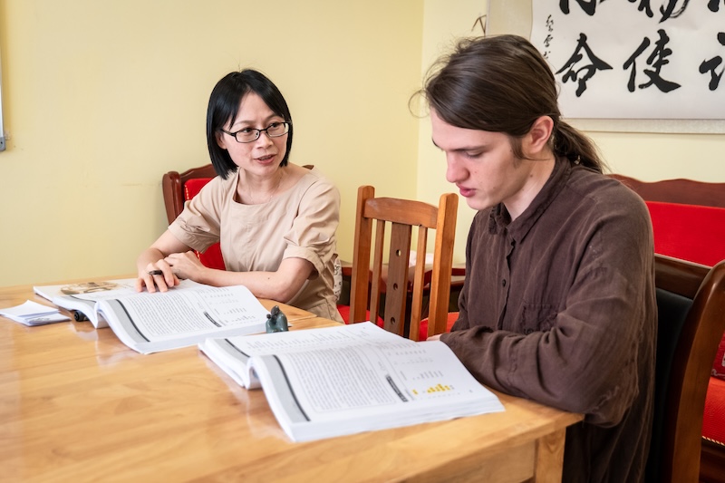 Un professeur chinois et un étudiant occidental sont assis côte à côte à une table en bois pour un cours particulier de mandarin. Leurs manuels sont ouverts devant eux ; le professeur désigne un passage et guide l’étudiant dans sa lecture. Une calligraphie chinoise orne le mur jaune derrière eux.