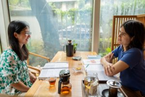 A teacher and student work through books at a wooden table by tall windows