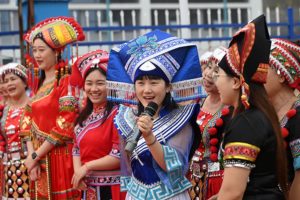 Women in colorful traditional dress and headdresses sing into a microphone