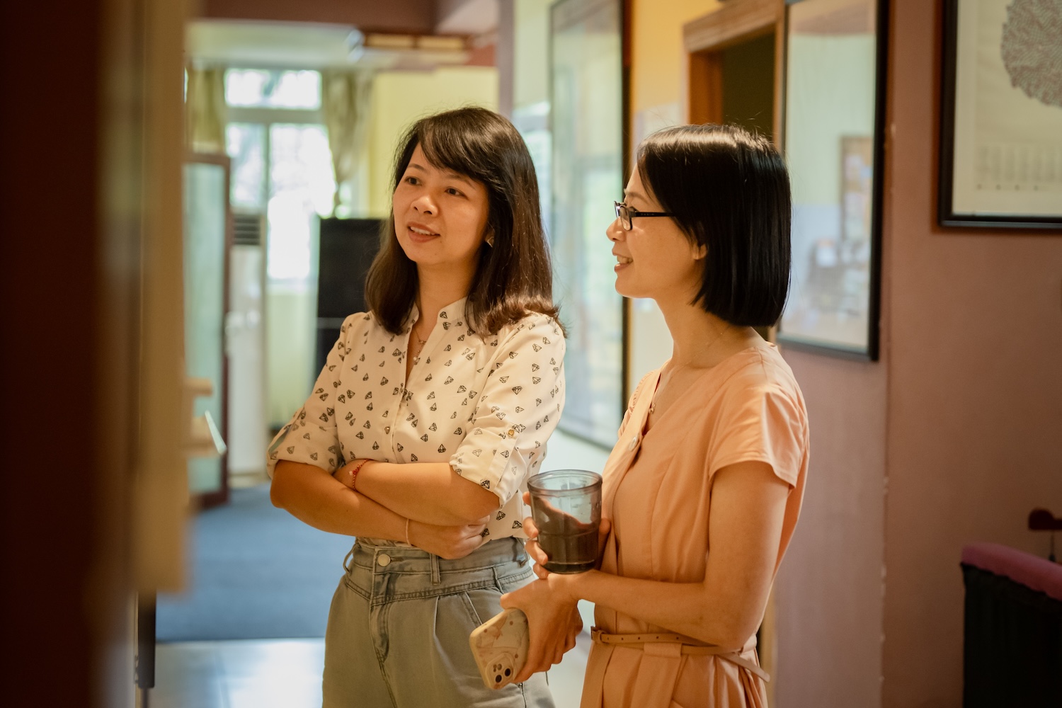 Two women standing in a softly lit hallway, talking and smiling; one wears a white patterned blouse and jeans with her arms crossed, while the other wears glasses and a light pink dress and holds a drink and a phone.
