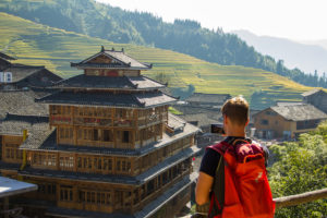 A traveler with a red backpack looks toward a wooden drum tower in a mountain village