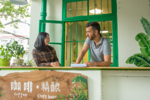 Two people talk across a cafe window counter surrounded by plants