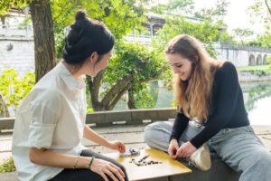 Two women play Go on a stone ledge beside a river, one sitting cross-legged