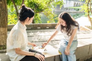 Two women play Go on a riverside ledge with an arched bridge behind them