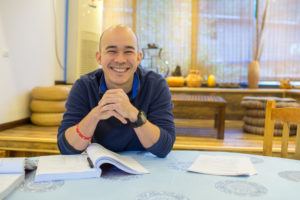 A smiling man sits at a table with an open textbook