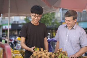 Two men smile while choosing longans at a fruit stand