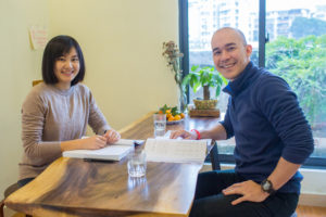 A teacher and student smile at a table beside a window
