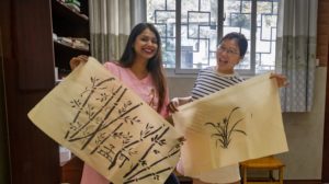 Two women hold up their finished ink paintings of bamboo and orchids