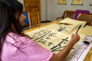 A woman in pink paints bamboo with an ink brush at a long table