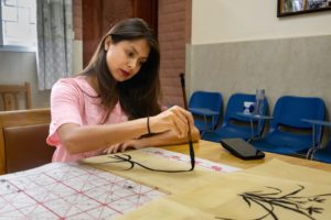 A woman paints ink bamboo on paper beside a row of blue chairs