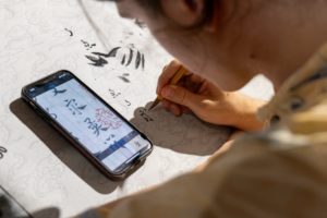A brush paints ink characters beside a phone showing a reference