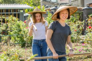 Two women in woven hats carry long wooden tools through a flower garden