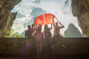 A group holds a red CLI banner overhead inside a cave opening against bright sky