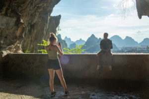 One person photographs another at a cave overlook facing karst peaks