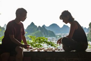Two people play Chinese chess in silhouette against karst peaks