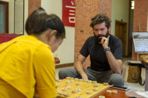 A woman in yellow and a man play Chinese chess near a 言 sign