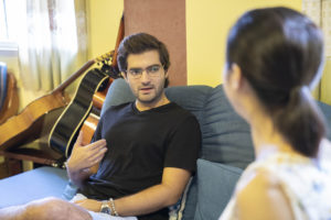 A man in glasses talks with a woman on a sofa, a guitar beside him