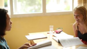 A smiling woman with an open textbook talks across a table by a sunny window