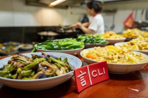 Plates of green beans and eggplant beside a CLI sign, a cook behind
