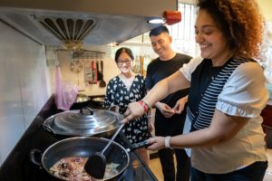 A woman stir-fries in a wok as two people watch and smile