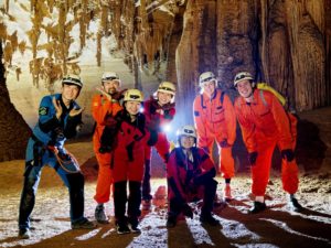 A group in caving suits and helmets poses inside a lit cave