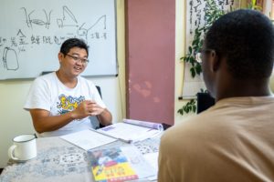 A smiling teacher sits at a table with notes and a whiteboard during a lesson