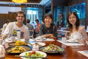 Three people smile at a restaurant table full of shared dishes