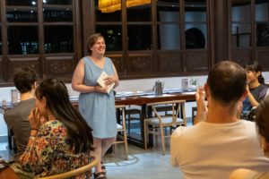 A woman holding a notebook speaks to people seated at restaurant tables