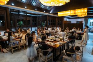 A large group dines at long tables in a restaurant with lantern lights